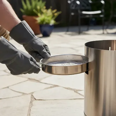 A person wearing protective gloves carefully removing the ash pan from a cooled Solo Stove Bonfire 2.0, showing the minimal ash residue and ease of cleaning.